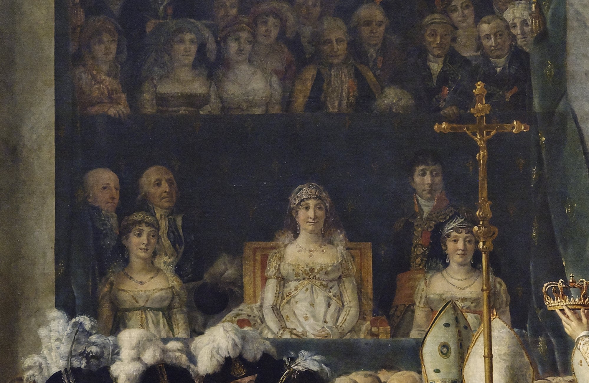 Detail of a raised box in which a woman in white with a diadem and veil sits on a red and gold chair, flanked by two younger women and two men, with a crowded gallery of spectators above.