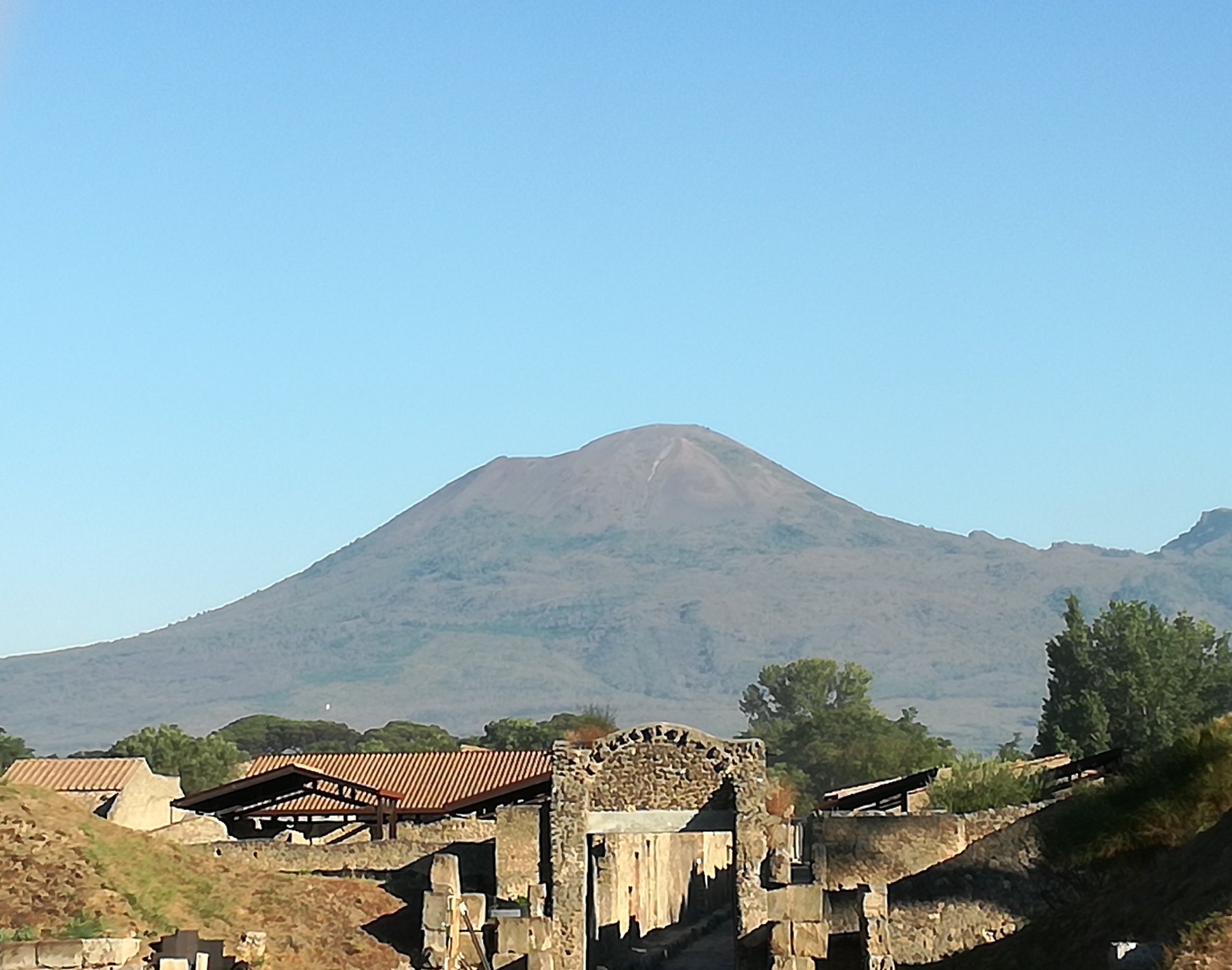 A view across the ruins of the Roman city of Pompeii with Mount Vesuvius rising in the background under a clear sky.