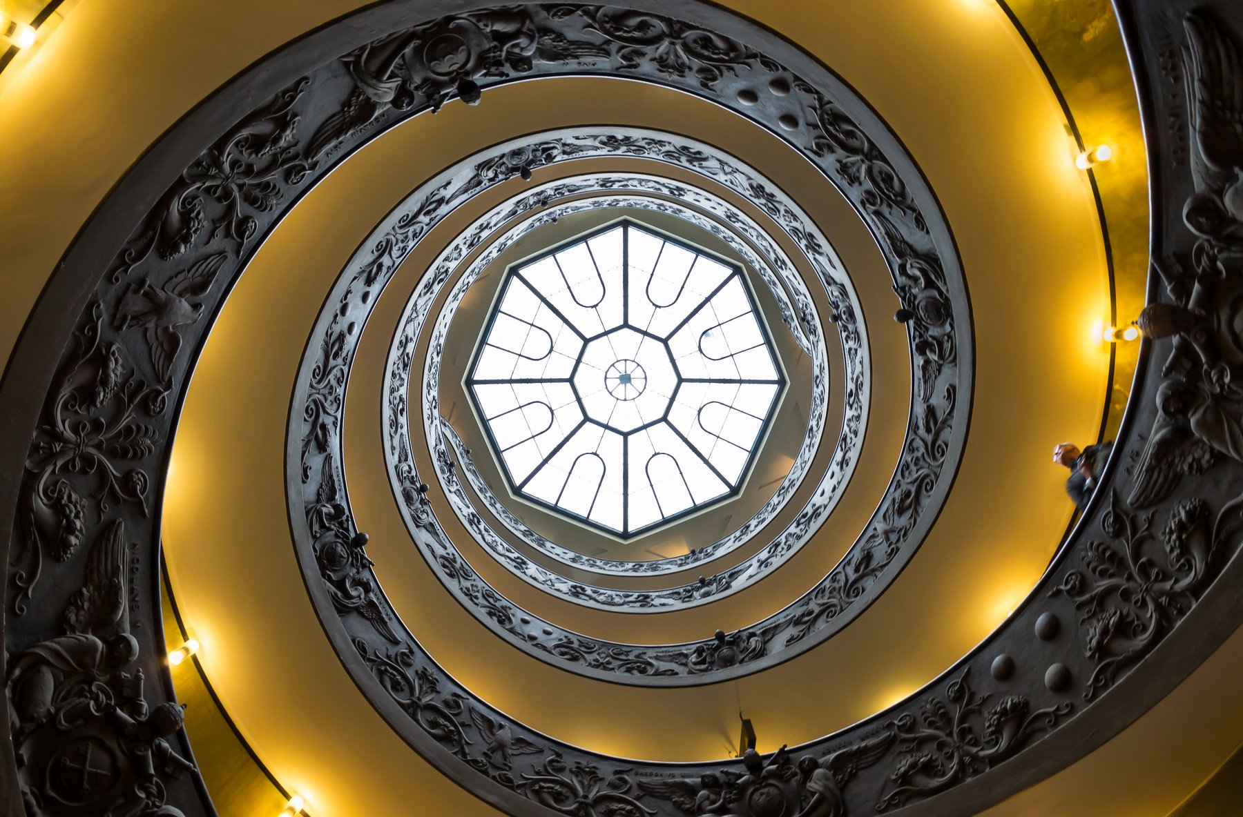 The double-helix spiral staircase at the exit of the Vatican Museums, designed by Giuseppe Momo in 1932, photographed looking up at the bronze handrail spiraling toward a stained-glass ceiling.